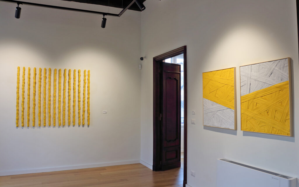 Veduta d'allestimento della mostra Fragili Sospensioni 2025 presso la Galleria Ellebi. Sulla parete destra è esposta l'opera "Dittico del giallo", composta da due quadri speculari caratterizzati da campiture geometriche diagonali bianche e gialle con fitte stratificazioni di materia e scrittura. Sulla parete sinistra dello sfondo è installata l'opera "Senza titolo giallo", una composizione lineare di 15 elementi verticali gialli avvolti da strisce di carta a spirale. Pavimento in legno chiaro. Opera realizzata dall'artista contemporaneo Giovanni Fava.
