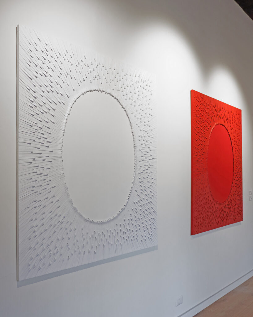 Scatto d'allestimento prospettico della mostra Fragili Sospensioni 2025 presso la Galleria Ellebi. In primo piano a sinistra è visibile l'opera "Grande silenzio bianco", una grande tela monocroma bianca caratterizzata da una fitta texture materica radiale in rilievo che converge verso un grande ovale liscio centrale. Sullo sfondo a destra è esposta la sua opera gemella monocroma di colore rosso dal titolo "Grande silenzio rosso". Opere realizzate dall'artista contemporaneo Giovanni Fava.