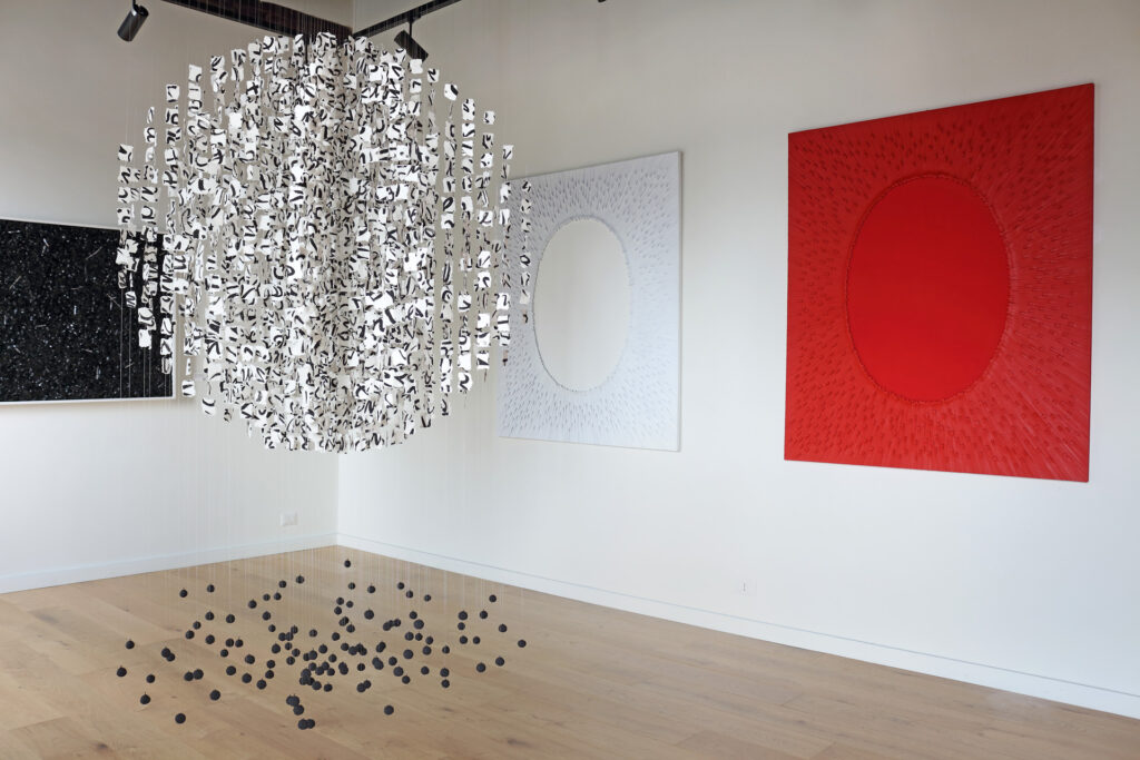 Veduta d'allestimento complessiva della mostra Fragili Sospensioni 2025 presso la Galleria Ellebi. In primo piano a sinistra è sospesa l'installazione omonima "Fragili sospensioni", una struttura geometrica sferica formata da moduli cartacei bianchi e neri che dialoga con piccoli elementi scuri a terra sul pavimento in legno chiaro. Sulla parete bianca di fondo sono esposte le opere monumentali monocrome con texture radiale in rilievo "Grande silenzio bianco" e "Grande silenzio rosso". Opera realizzata dall'artista contemporaneo Giovanni Fava.