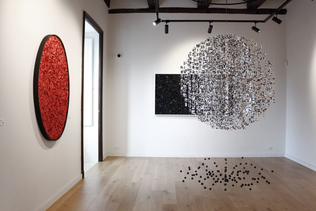 Veduta d'allestimento della mostra Fragili Sospensioni 2025 presso la Galleria Ellebi. In primo piano un'installazione sferica sospesa formata da moduli cartacei bianchi e neri. Sulle pareti bianche sono esposte un'opera circolare rossa e, sullo sfondo, un'opera rettangolare nera. Pavimento in legno chiaro. Opera realizzata dall'artista contemporaneo Giovanni Fava.