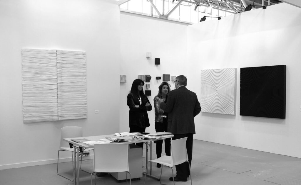 Fotografia in bianco e nero documentaria dell'allestimento di uno stand espositivo fieristico. All'interno dello spazio geometrico, tre figure umane sono disposte in piedi al centro accanto a un tavolo tecnico con cataloghi. Sulla parete sinistra è installato un grande dittico bianco rettangolare a rilievo plastico formato da fitte stratificazioni cartacee a andamento orizzontale. Sulla parete di fondo sono disposti due grandi pannelli quadrati speculari, uno bianco e uno nero, caratterizzati da una texture a rilievo a cerchi concentrici con fulcro centrale. Struttura industriale del soffitto con fari visibile in alto. Documentazione del progetto espositivo di Giovanni Fava.