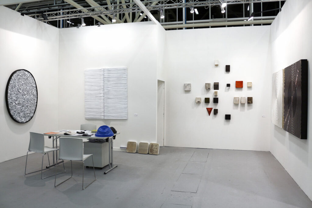 otografia a colori documentaria di uno stand espositivo d'arte contemporanea con pareti bianche e pavimento grigio neutro. Sulla parete sinistra sono installate un'opera ellittica con cornice nera e fitta texture cartacea chiara a rilievo e un dittico rettangolare bianco a linee orizzontali stratificate. Sulla parete di fondo è disposta una costellazione geometrica di piccoli blocchi scultorei polimaterici. Sulla parete destra si articola una serie di tre grandi pannelli quadrati accostati, caratterizzati da superfici lavorate a cerchi concentrici lucidi e opachi neri e bianchi. Un tavolo tecnico con sedie bianche e un casco blu si posiziona sulla sinistra. Allestimento di Giovanni Fava.