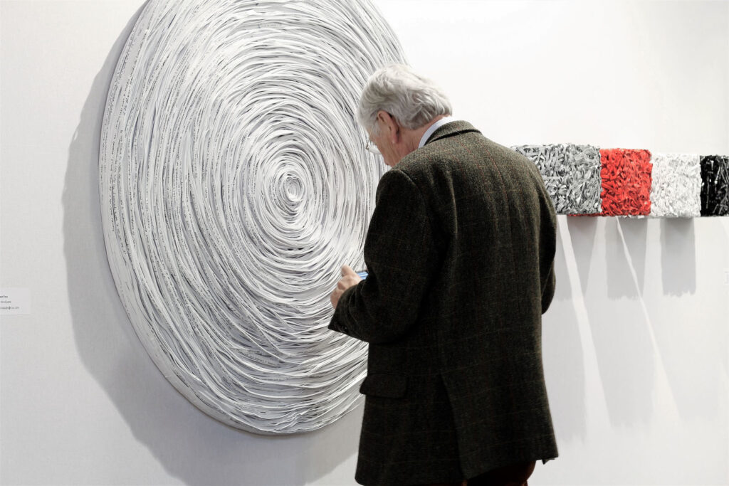 Veduta d'allestimento a YIA Art Fair Maastricht 2017 con visitatore di spalle in osservazione. In primo piano, opera plastica circolare monumentale a parete con fitta texture radiale e concentrica in rilievo formata da stratificazioni di carta bianca. Sullo sfondo a destra, sequenza lineare e geometrica di sculture volumetriche a forma di cubo con superfici materiche composte da frammenti di supporti cartacei triturati e stratificati in tonalità grigio, rosso, bianco e nero. Opere di Giovanni Fava.