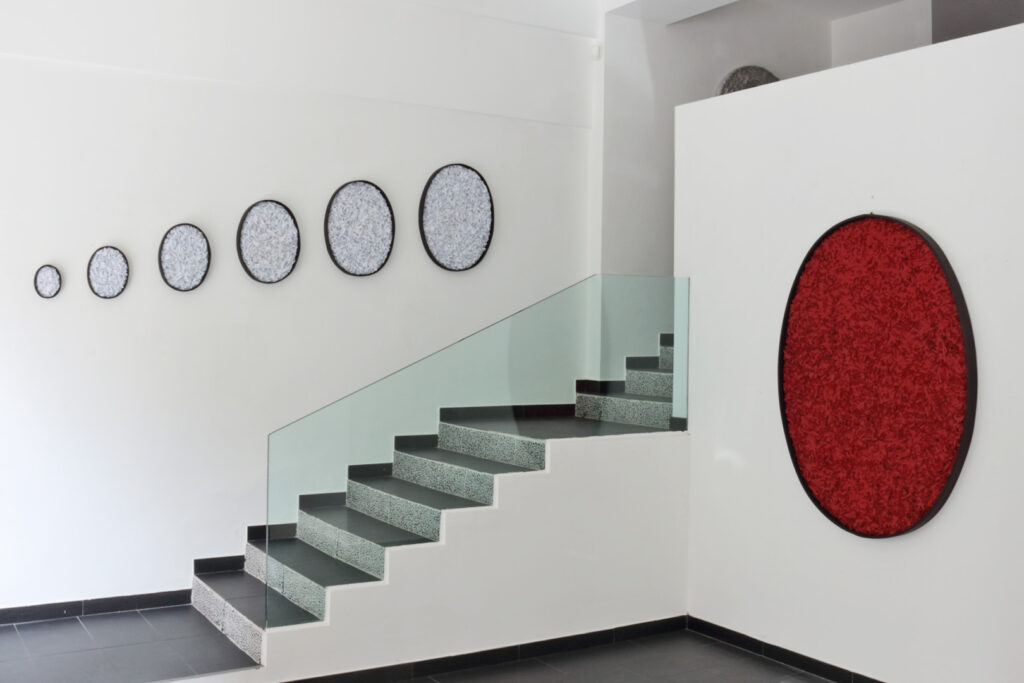 Spazio espositivo interno con scala in pietra grigio scuro e parapetto in lastre di vetro trasparente. Parete sinistra bianca con installazione seriale di sei opere di forma ovale disposte lungo una linea diagonale in progressione dimensionale crescente da sinistra a destra; superficie interna a fitta texture frammentata bianco e grigio con cornici perimetrali nere. Parete destra avanzata con grande opera monumentale di forma ovale monocroma rosso saturo a forte rilievo plastico e densità materica compressa entro cornice nera. Pavimentazione scura uniforme. Documentazione d'arte contemporanea di Giovanni Fava.