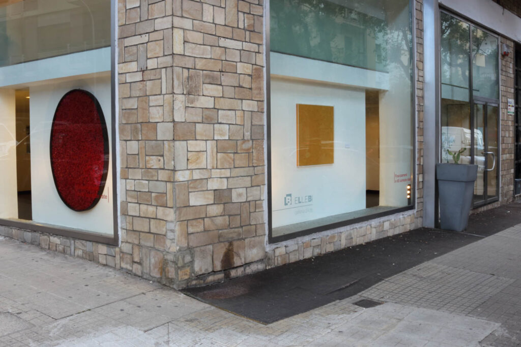 Inquadratura esterna d'angolo di architettura espositiva urbana. Facciata in blocchi di pietra squadrata chiara con ampie vetrate trasparenti terra-cielo. Attraverso la vetrata sinistra, visione di grande opera ovale monocroma rossa ad alto rilievo plastico e densità materica compressa. Attraverso la vetrata frontale destra, esposizione di pannello quadrangolare monocromo giallo saturo su parete bianca retrostante con illuminazione direzionale. Logotipo istituzionale Galleria Ellebi impresso su vetro. Marciapiede in cemento in primo piano. Documentazione d'arte contemporanea di Giovanni Fava.