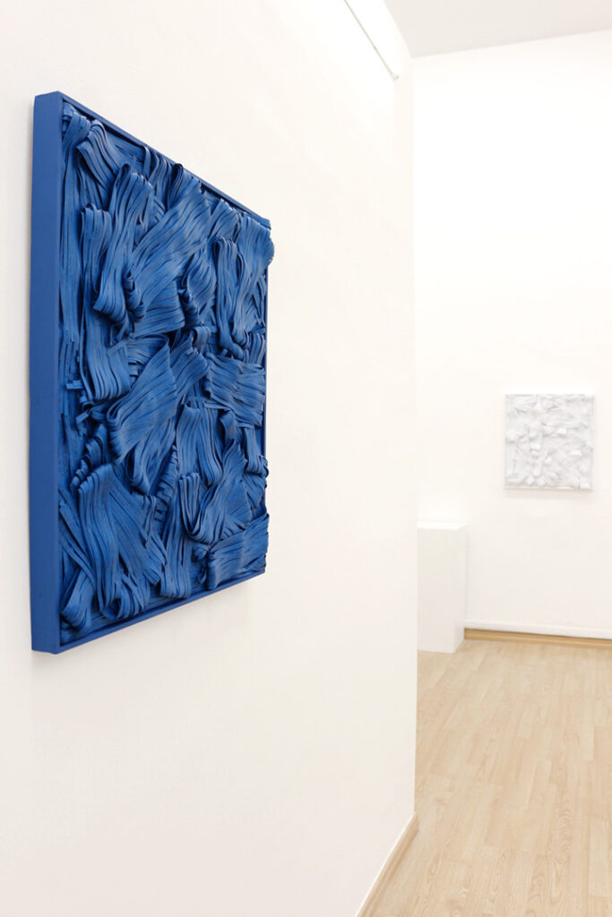 Inquadratura prospettica angolare e radente di parete espositiva bianca. In primo piano a sinistra, sezione laterale di pannello quadrangolare monocromo blu saturo con fitta stratificazione di bande materiche sinuose e ripiegate ad alto spessore plastico. Sullo sfondo in asse prospettico, secondo pannello quadrangolare a fondo chiaro con tessitura materica irregolare a rilievo attenuato. Pavimentazione lignea chiara a sviluppo diagonale. Documentazione d'arte contemporanea di Giovanni Fava.