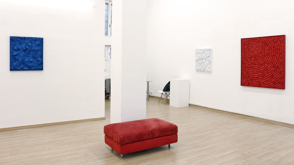 Spazio espositivo minimale. Pareti bianche e pavimentazione lignea chiara. Parete sinistra avanzata con pannello quadrangolare monocromo blu a stesure materiche sbalzate ad alto rilievo con forti contrasti chiaroscurali. Colonna portante bianca a sezione verticale al centro. Parete di fondo con opera quadrata a fondo bianco e stratificazioni materiche irregolari. Parete destra con grande pannello quadrangolare monocromo rosso a densa struttura lineare a spirale concentrica continua. Primo piano con volume parallelepipedo imbottito orizzontale rosso cupo a terra. Documentazione d'arte contemporanea di Giovanni Fava.