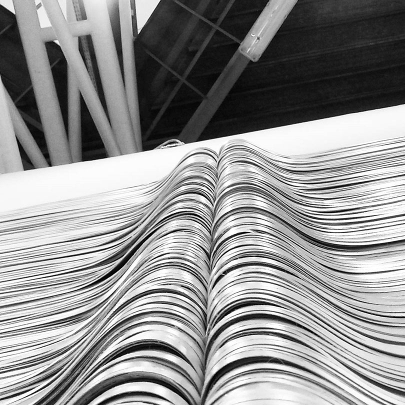 Fotografia in bianco e nero incentrata sul dettaglio macro di una struttura plastica speculare. La superficie è costituita da una sequenza fitta, parallela e stratificata di bande e strisce cartacee curvate a formare due volumi simmetrici a cresta ondulata. L'andamento delle linee parallele genera una modulazione geometrica regolare con flussi chiaroscurali netti determinati dall'incidenza della luce sulle curvature dei supporti. Elementi tubolari metallici visibili sullo sfondo superiore. Opera di Giovanni Fava.