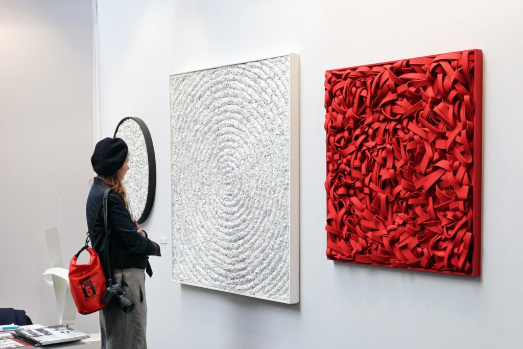 Veduta prospettica dello stand espositivo della Galleria Ellebi a YIA Art Fair Parigi 2017 con visitatrice di profilo dotata di macchina fotografica. A parete sono installate un'opera bianca di formato quadrangolare a rilievo materico con fitti cerchi concentrici e, a destra, un'opera rossa a tecnica mista su carta caratterizzata da una complessa stratificazione di strisce e matasse cartacee volumetriche ad alto contrasto. Opere di Giovanni Fava.
