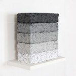 Another word in the brick 2021, opera d'arte contemporanea, tecnica mista su carta e legno. Cinque mattoni con texture di pensieri su carta triturati e compressi poggiano su una mensola bianca. Gli elementi partono dall'alto con tonalità grigio scuro fino a sfumare al bianco nell'elemento in basso. Opera realizzata dall'artista Giovanni Fava.