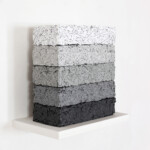 Another word in the brick 2021, opera d'arte contemporanea, tecnica mista su carta e legno. Cinque mattoni con texture di pensieri su carta triturati e compressi poggiano su una mensola bianca. Gli elementi partono dal basso con tonalità grigio scuro fino a sfumare al bianco nell'elemento in alto. Opera realizzata dall'artista Giovanni Fava.