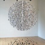 Fragili sospensioni 2025, installazione d'arte contemporanea, tecnica mista su carta, fili di nylon, alluminio e sabbia. Composta da un nucleo centrale a forma di sfera formata da tantissimi frammenti di carta con tracce di scrittura, sospesa nel vuoto attraverso dei fili di nylon. Nella parte bassa, per ogni filo, un nucleo di piccoli pesi dalla forma sferica di colore nero sospesi come un universo di piccoli pianeti. Opera realizzata dall'artista Giovanni Fava.